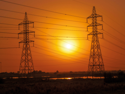 Tendido eléctrico entre dos torretas sobre un fondo-paisaje de atardecer. El Sol, entre ambas siluetas, ilumina un cielo anaranjando
